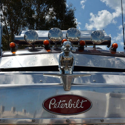 Sidabrinis sunkvežimio priekis su apvaliais žibintais ir raudonu „Peterbilt“ logotipu.