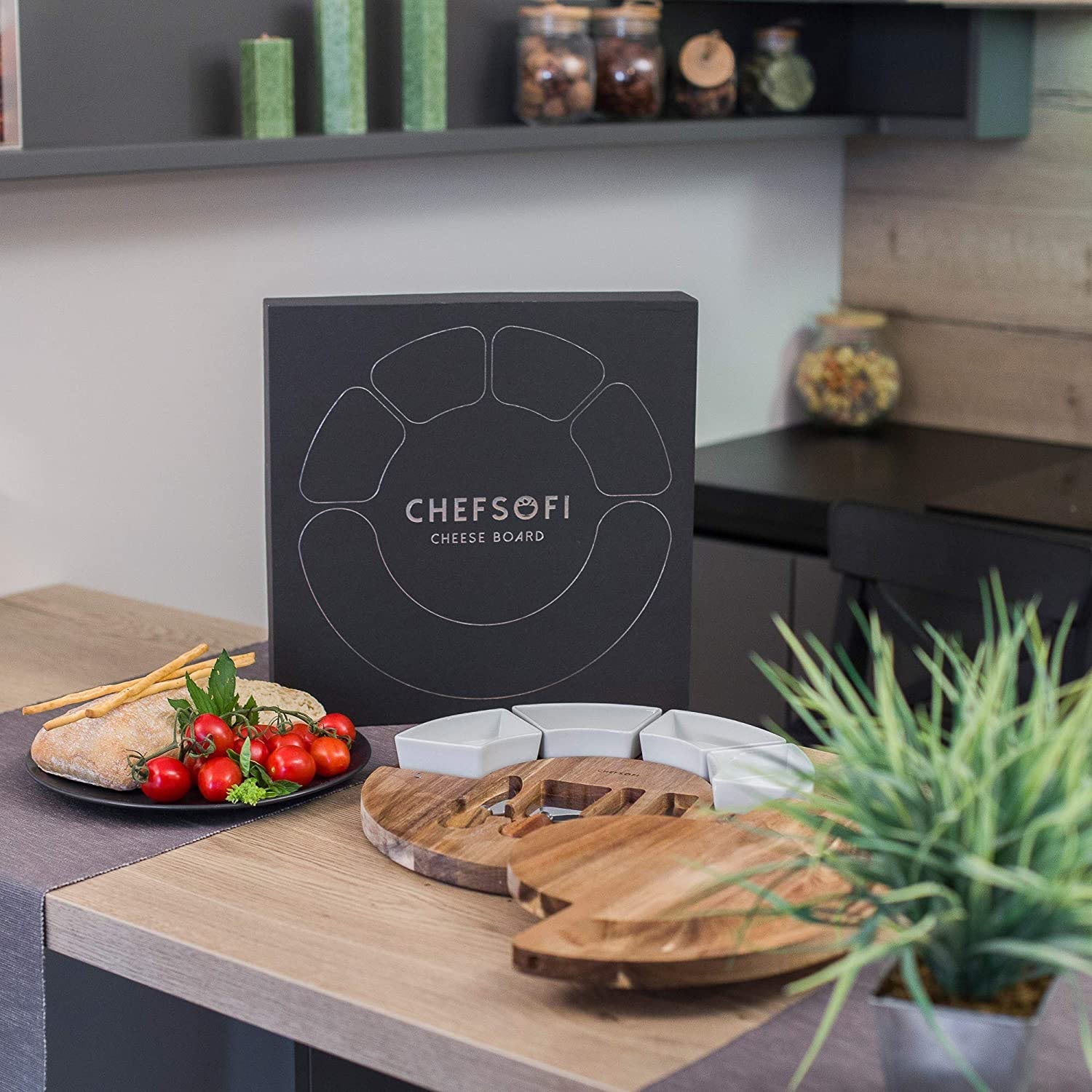 Juodas sūrių padėklas su baltomis lėkštelėmis, medinėmis detalėmis ir šviežiais ingredientais.