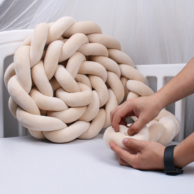 Šviesiai kreminės spalvos, minkštas, pintas pagalvės dizainas su apvaliais formos elementais.