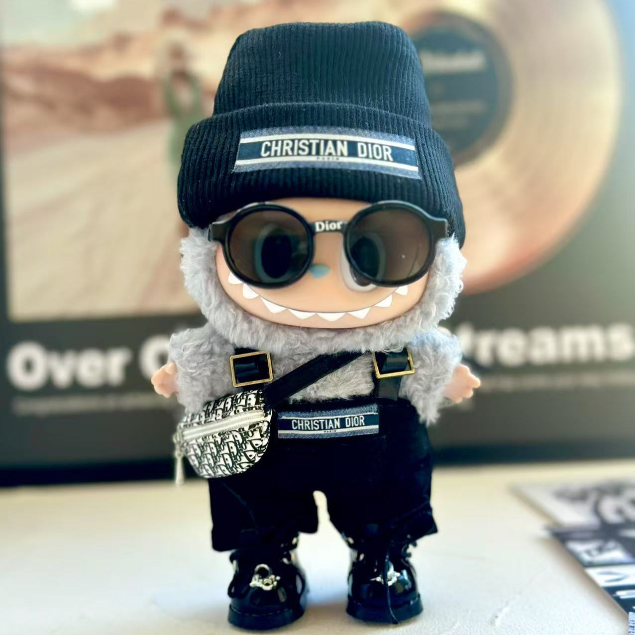 Labubu lėlė, apvalus veidas, juoda kepurė, pilkas pūkuotas kailiukas, blizgūs juodi batai