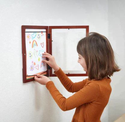 Mergaitė deda piešinį į atvertą rudą sieninį rėmelį